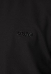 Sort stof med en struktureret finish, der har et præget "HUGO"-logo i en matchende farve, placeret på venstre side.