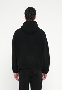 Čierna, veľká mikina s kapucňou vyrobená z mäkkej tkaniny, obsahujúca veľkú kapucňu, voľný strih a rebrované manžety. Textúrovaný povrch bez viditeľných log.