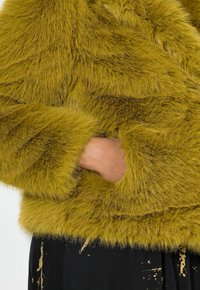 Faux furjakke i olivengrøn med en blød, fluffy tekstur, der har lange ærmer og en sidelomme. Sort stof med gyldne accenter er synligt nedenfor.