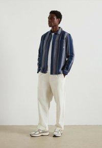 Niebieska i kremowa koszula z guzikami w paski, biała koszulka pod spodem, kremowe szerokie spodnie i szare sneakersy z ciemnymi akcentami. Gładka tekstura.