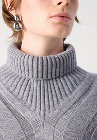 Sivko pleteno jopico z visokim, rebrastim turtleneck-om. Ima teksturiran vzorec v obliki chevrona in mehak volneni material. Vidne so sijoče viseče uhane.