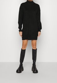 Robe noire en maille à manches longues et col montant, associée à des bottes noires à plateforme montant jusqu'aux genoux, créant une silhouette élancée.