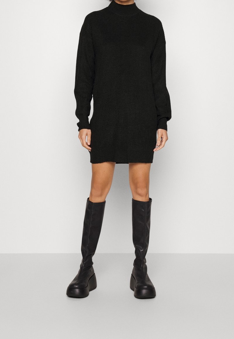 Robe noire en maille à manches longues et col montant, associée à des bottes noires à plateforme montant jusqu'aux genoux, créant une silhouette élancée.