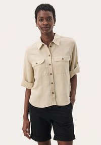 Camicia beige con bottoni e due tasche sul petto, maniche arrotolabili e orlo curvo. Indossata con pantaloni corti neri, caratterizzati da una consistenza morbida.