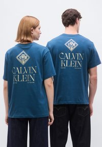 Μπλε βαμβακερά μπλουζάκια με γραφικό στο πίσω μέρος; διαθέτουν το "CALVIN KLEIN" με χρυσά γράμματα και ένα λουλουδένιο έμβλημα μέσα σε διαμάντι.