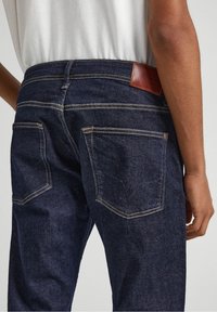 Jeans de mezclilla azul oscuro con dos bolsillos traseros con acentos cosidos y un parche de cuero marrón en la cintura. Textura suave, diseño ajustado.