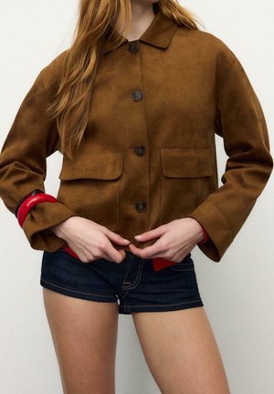 Mujer con una chaqueta marrón de ante con botones y bolsillos con solapa, y pantalones cortos vaqueros oscuros, ajustándose la parte inferior de la chaqueta con ambas manos.