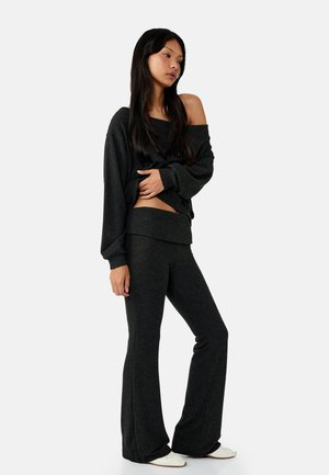 Kobieta w ciemnym swetrze z odkrytymi ramionami i dopasowanych rozkloszowanych spodniach, stoi z jedną ręką na talii, nosi białe buty.