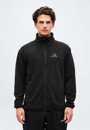 Giovane uomo che indossa una giacca in pile nera Adidas con zip, mani rilassate lungo i fianchi, rivolto verso davanti su uno sfondo chiaro e semplice.