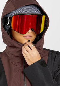 Skijacke mit einer braunen Kapuze, schwarzem Körper und strukturiertem Stoff. Verfügt über große rotgetönte Skibrille und eine weiche Innenauskleidung.