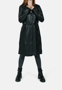 Gabardina negra de cuero con cintura ceñida, mangas largas y cuello con muesca, combinada con leggings oscuros a rayas y botas negras de combate.