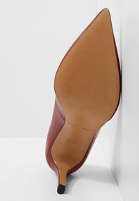 Chaussure à talon haut en cuir rouge à bout pointu vue de la semelle avec un talon stiletto étroit appuyé contre un mur blanc d'angle.