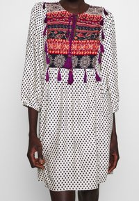 Kvinna som bär en vit klänning med svarta mönster, med en färgglad broderad bröstpanel och lila tofsar, med trekvartsärmar.