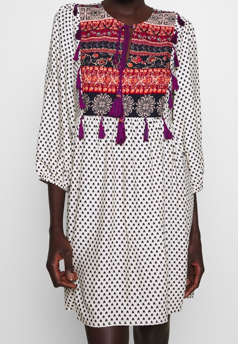 Femme portant une robe blanche avec des motifs noirs, dotée d'un panneau de poitrine brodé coloré et de pompons violets, avec des manches trois quarts.