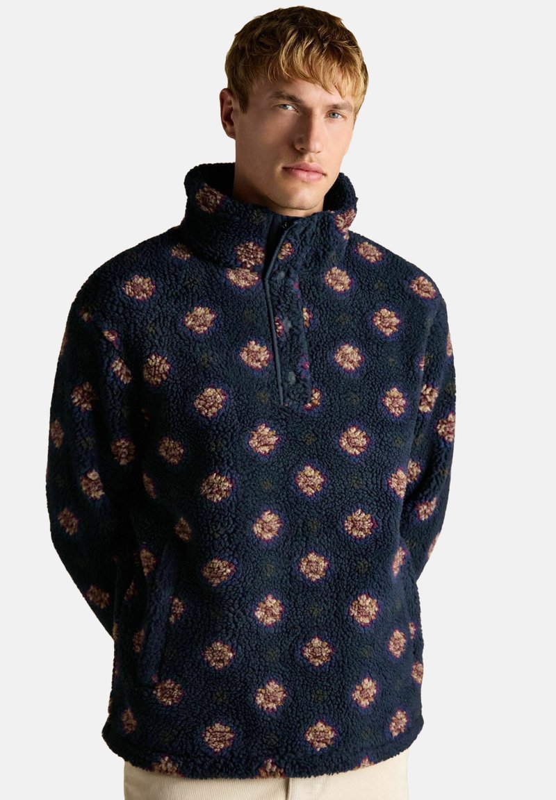 Fleece-Pullover mit hohem Kragen, mit marineblauem Grund und floralem Muster in Beige und Lila, versehen mit einem kurzen Reißverschluss.