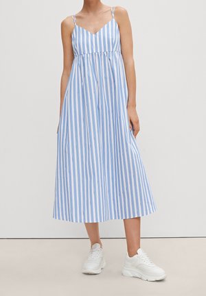 Mujer vestida con un vestido midi sin mangas con rayas verticales azules y blancas y zapatillas blancas gruesas, posando frente a un fondo liso.