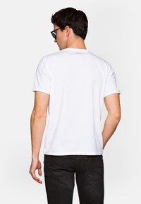 Biała bawełniana koszulka z krótkim rękawem, okrągłym dekoltem i małym logo nadrukowanym na plecach. Noszona z czarnymi dżinsami.