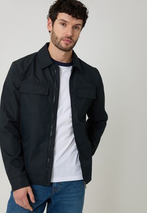 Mand med mørkt krøllet hår iført en sort lynlåstrøje over en hvid t-shirt og blå jeans, stående mod en ensfarvet lysgrå baggrund.