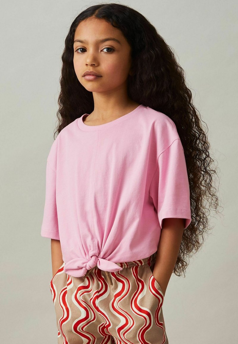Chica con cabello rizado largo que lleva una camiseta corta rosa con nudos y pantalones beige con rayas rojas en forma de onda, con las manos en los bolsillos.