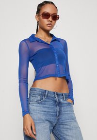Blue sheer long-sleeve shirt with a collar, button front, and cropped design, paired with light blue denim jeans.