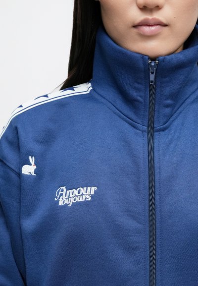 Tmavomodrá bunda na zips s vysokým golierom, so štýlovými bielymi hviezdičkami na pleciach, vyšívaným zajacím logom a nápisom „Amour toujours“.