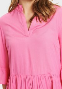 Rosa Bluse mit V-Ausschnitt, dreiviertel Ärmeln und geraffter Taille. Glatter Stoff mit weicher Textur und dezentem Glanz.