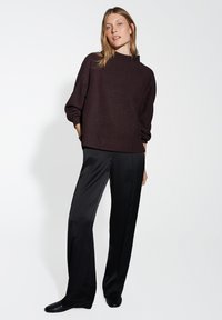 Maglione marrone con colletto alto, vestibilità rilassata e spalle scese abbinato a pantaloni neri a gamba larga e scarpe nere su uno sfondo bianco.