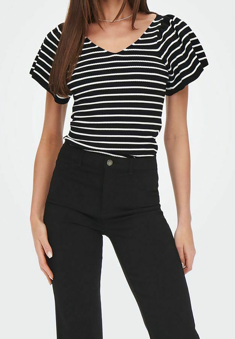Musta-valkoraidallinen paita, jossa on röyhelöhihat, V-pääntie ja resoripintainen rakenne. Yhdistetty korkeavyötäröisiin mustiin housuihin, joissa on nappi.