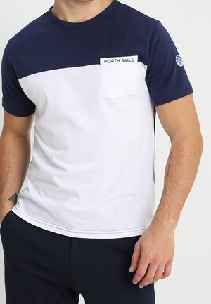 Homme portant un t-shirt bicolore bleu marine et blanc avec une poche poitrine blanche libellée « North Sails » et un pantalon bleu marine.