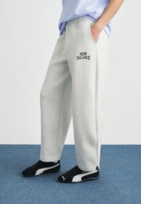 Ανοιχτόγριμη φόρμα με άνετη εφαρμογή, διαθέτει πλαϊνές τσέπες και κεντημένο μαύρο λογότυπο "New Balance". Συνδυάζεται με μαύρα αθλητικά παπούτσια.