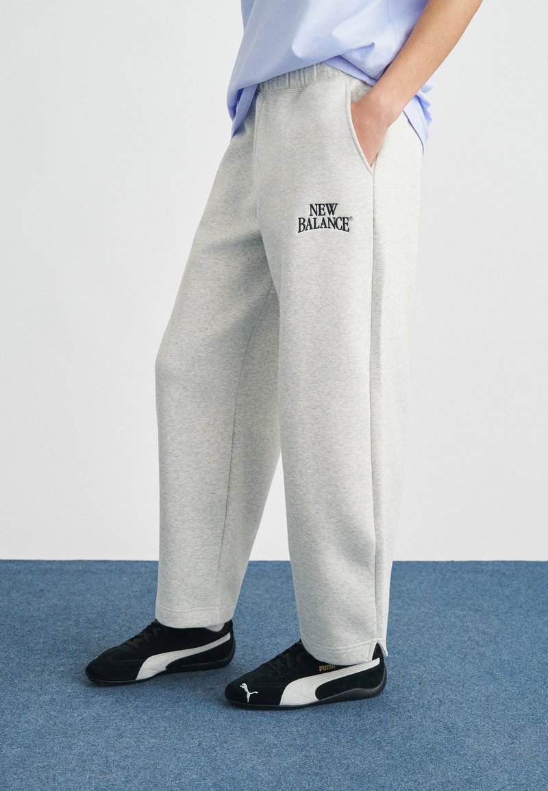 Ανοιχτόγριμη φόρμα με άνετη εφαρμογή, διαθέτει πλαϊνές τσέπες και κεντημένο μαύρο λογότυπο "New Balance". Συνδυάζεται με μαύρα αθλητικά παπούτσια.