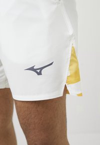 Detailaufnahme einer Person, die weiße Sportshorts mit einem schwarzen Logo und gelb gemusterten Details an der Seite trägt.