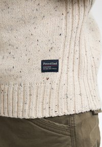 Maglione lavorato a maglia color crema con orlo a coste, caratterizzato da scaglie multicolori e un'etichetta nera con "Petrol Ind" vicino al fondo.