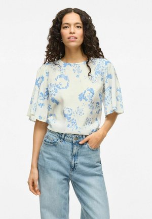 Lätt, krämfärgad blus med blå blommönster, korta volangärmar och rund halsringning; matchad med högmidjade ljusblå jeans.