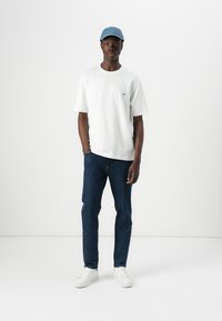 White cotton t-shirt, blue denim jeans, and white sneakers; blue cap with logo; model stands against a plain white background.