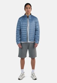 Ljusblå, vadderad pufferjacka med stående krage; kombinerad med en grå t-shirt och grå shorts; vita sneakers och strumpor fullbordar looken.