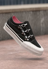 Zapatilla negra con detalles de hebillas metálicas, suela blanca estriada y forro interior rosa. Presenta una textura suave y un diseño moderno.
