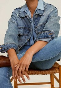 Chaqueta de mezclilla con un cuello azul claro, detalles desgastados y grandes bolsillos frontales. Usada sobre una camisa azul clara y combinada con jeans.