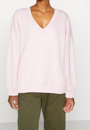 Persoon die een lichtroze V-hals lange mouwen sweatshirt met subtiel logo en olijfgroene broek draagt, getoond van nek tot midden dijbeen.