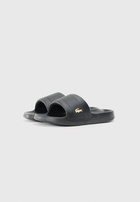 Svart dunsandaler laget av syntetisk materiale med to brede stropper, teksturert design og en gull Lacoste-logo på en av stroppene.