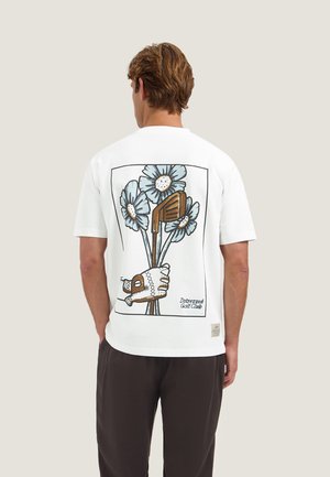Man draagt een wit t-shirt met afbeelding van een golfhandschoen die clubstelen met blauwe bloemen vasthoudt, staand voor een effen achtergrond.