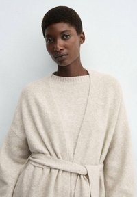 Cardigan avvolgente in lana beige con un ampio colletto e chiusura a cravatta in vita, dotato di maniche larghe e una finitura morbida e testurizzata.