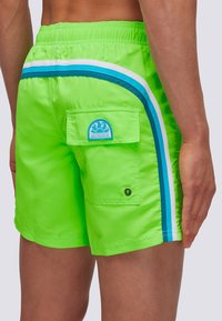 I pantaloni da bagno verde neon presentano un motivo a strisce bianche e blu sui lati, una tasca sul retro con un'etichetta del logo e una cintura elastica.
