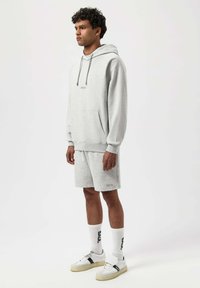 Hoodie harmaa, jossa on nauhat ja etutasku, yhdistettynä yhteensopiviin shortseihin. Koristeltu logolla ja valkoisilla sukilla, joissa on brändäys. Rentoa jalkineita täydentää ilmeen.