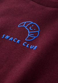 Cruasán sonriente bordado en azul y texto "SNACK CLUB" sobre tela granate.