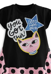 T-shirt noir à manches courtes avec un graphique de chat rose portant des lunettes et le texte "Tu peux le faire !" et "QUE DES BONNES VIBRATIONS" en bleu. Pois roses en bas.