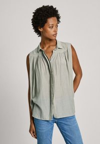 Blusa senza maniche verde chiaro con una chiusura a bottoni, dettagli plissettati e colletto. Abbinata a jeans blu, con una vestibilità drappeggiata.