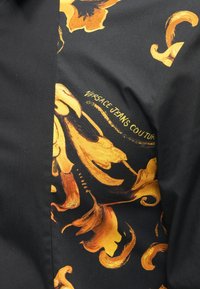 Črna srajca iz tkanine s srebrno in oranžno cvetličnim vzorcem na eni strani; vključuje natisnjeno blagovno znamko "Versace Jeans Couture".