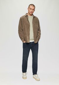 Brązowa kurtka z kordurowej tkaniny z kieszenią, zielony sweter pod spodem, ciemne dżinsy i białe trampki z beżowymi akcentami.