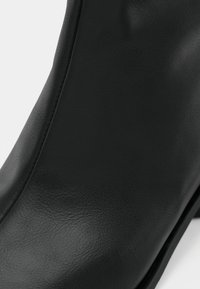 Stivale alla caviglia in pelle nera con texture liscia, caratterizzato da una forma elegante e cuciture lungo i bordi. Design minimalista senza hardware visibile.